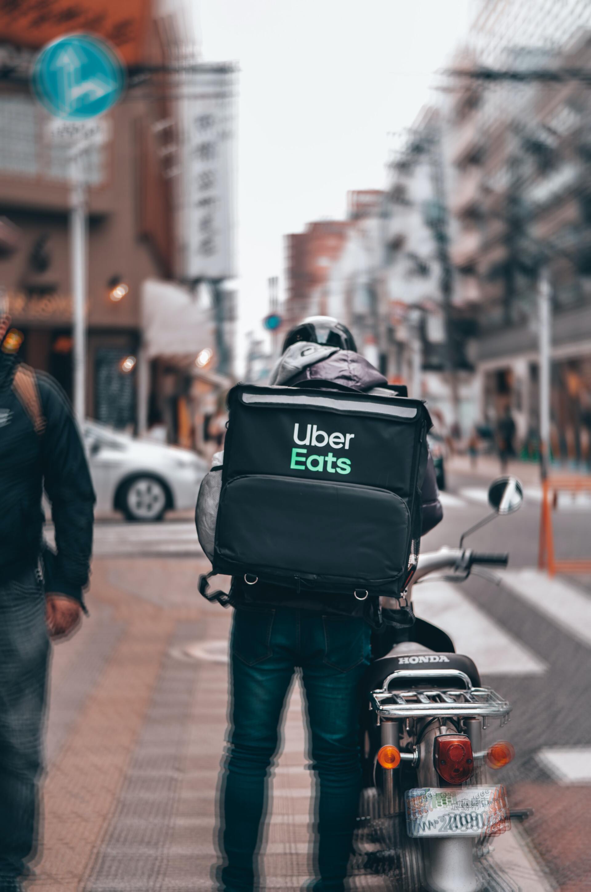 Uber Eats delivery person on a motorcycle in a city, wearing a backpack, waiting at a crosswalk.