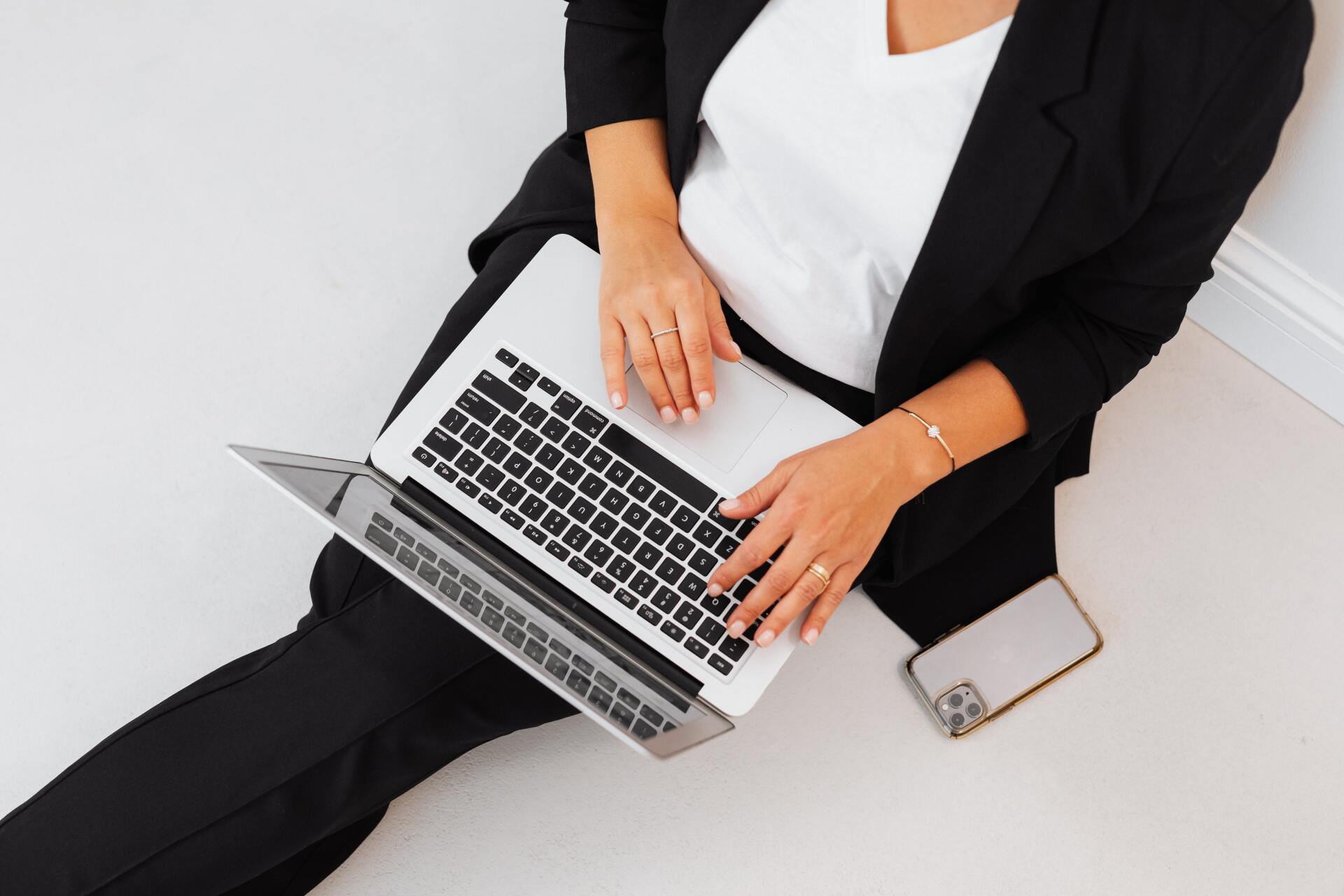 Uma pessoa vestindo um blazer preto está sentada em um piso claro, digitando em um laptop com um smartphone ao lado.