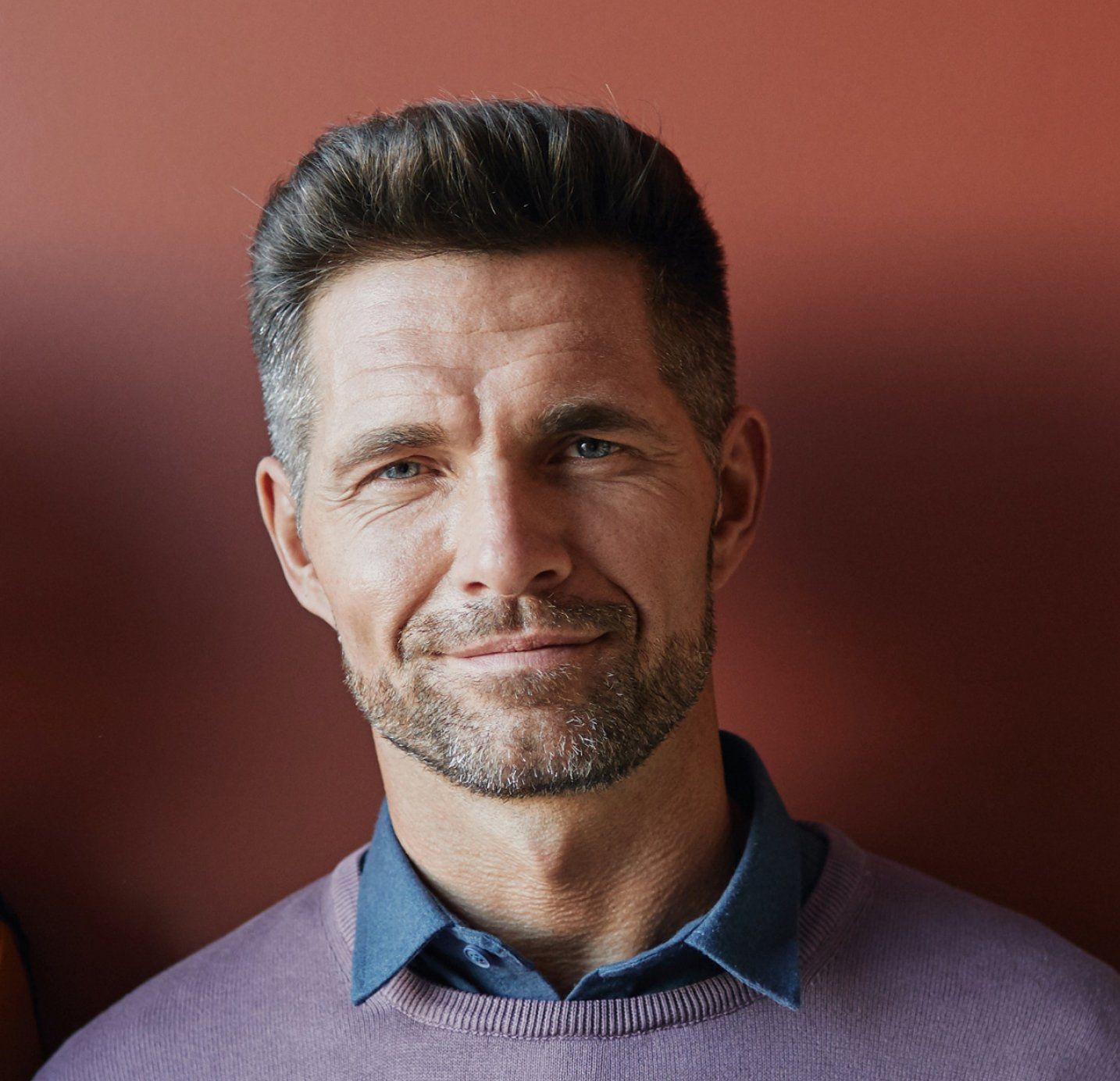 Man with short dark hair and a trimmed beard smiles, wearing a purple sweater over a blue collared shirt.