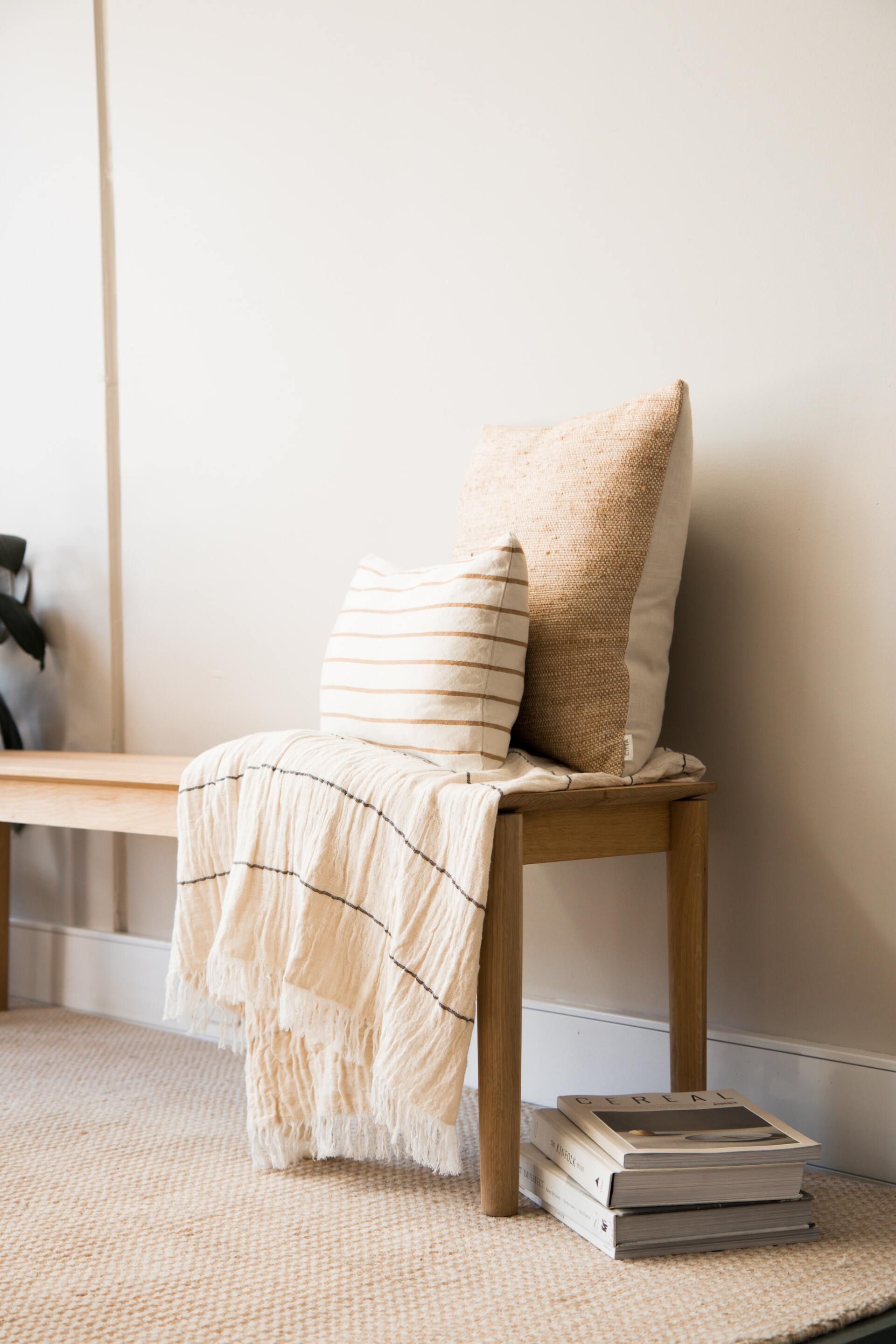 Wooden bench with pillows and a blanket, beside a stack of books on a rug.