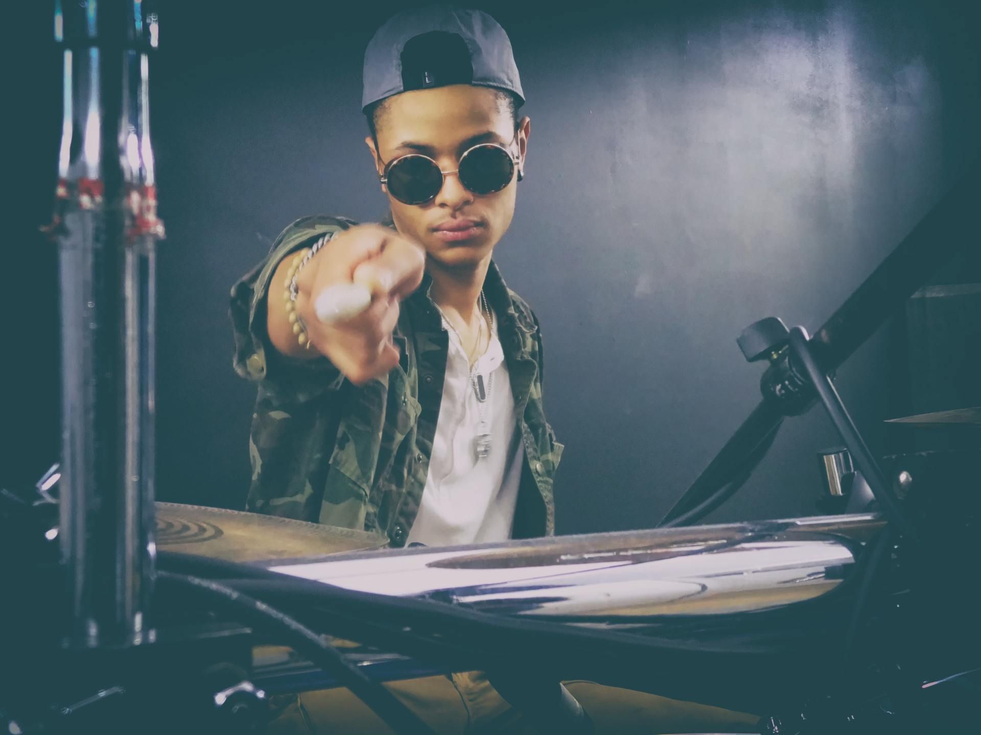 DJ at turntables pointing at the camera in a dim studio, wearing sunglasses and a cap
