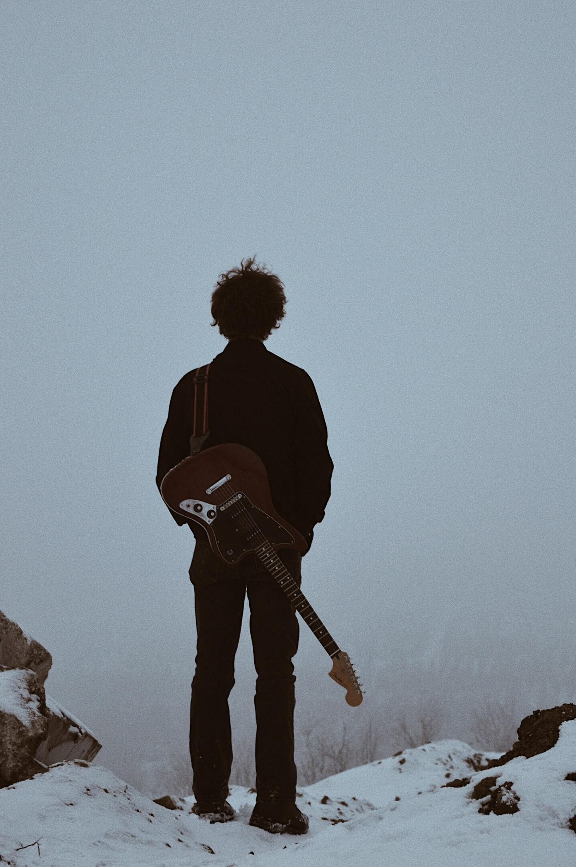 Silhouetted person stands on snowy rocks at dusk, holding a guitar against a pale sky.