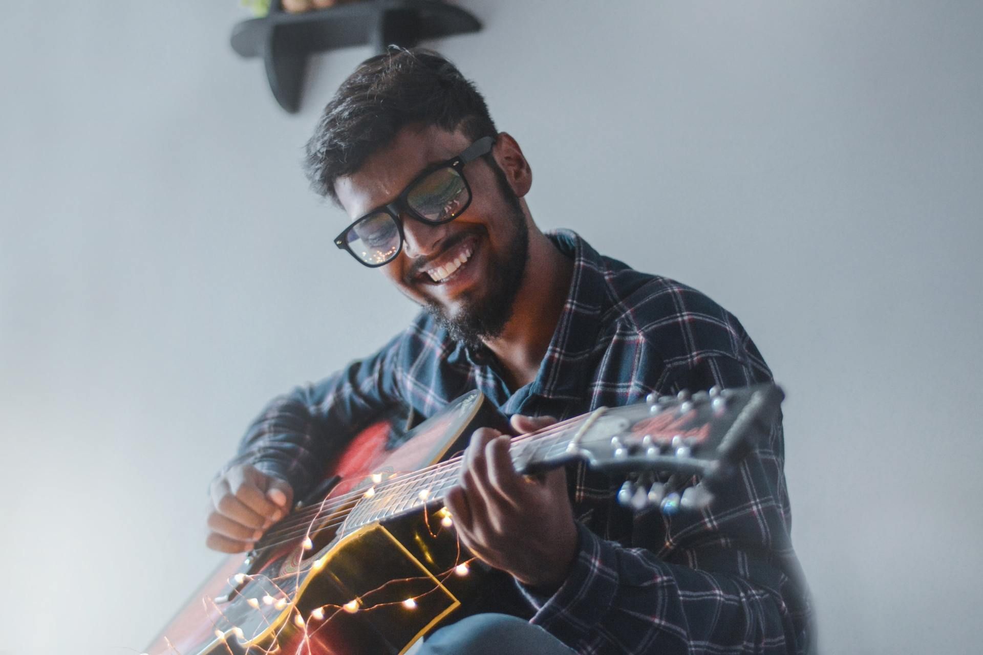 Smiling man in glasses playing an acoustic guitar indoors with warm string lights nearby