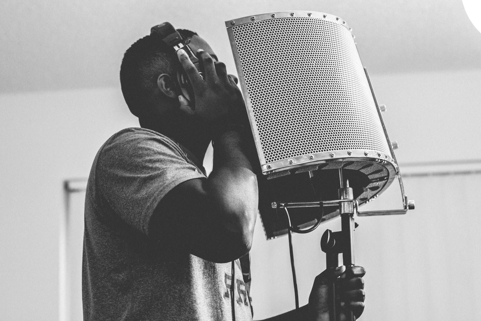 Person singing into a microphone in a recording booth, wearing headphones and a T-shirt.