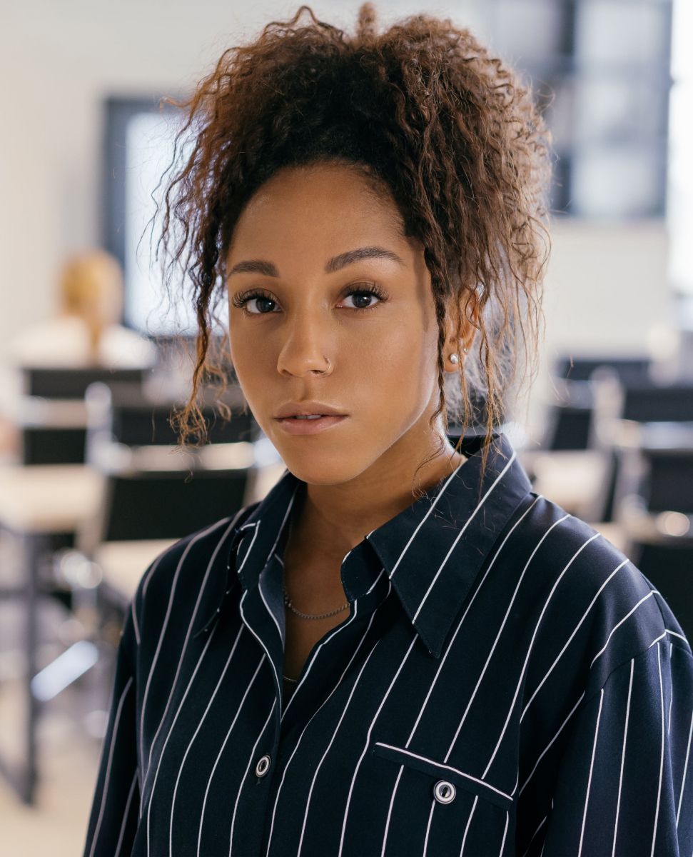 Woman with curly brown hair, wearing a striped shirt, looking directly at the camera in a classroom setting.