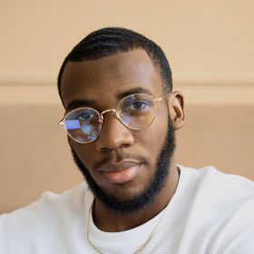 Young Black man with short waves, wearing round gold glasses and a white shirt, looking at the camera.