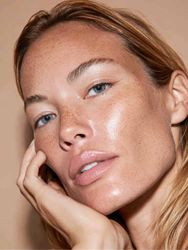 Close-up portrait of a person with dewy skin, resting their face on their hand against a beige background