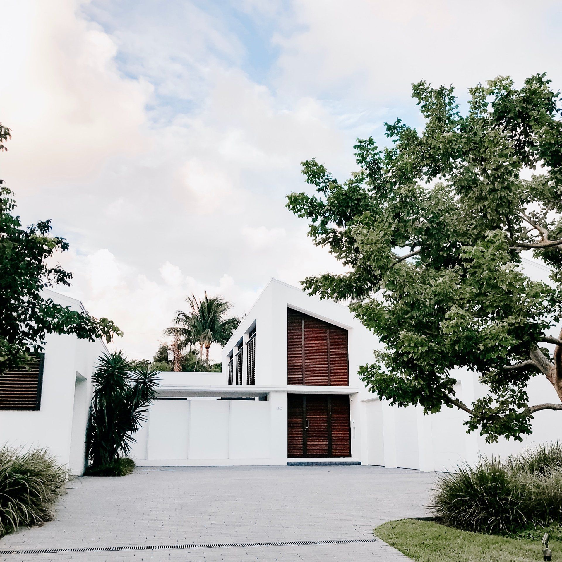 Modern white house with a sloped roof, large windows, and trees framing a driveway under a cloudy sky