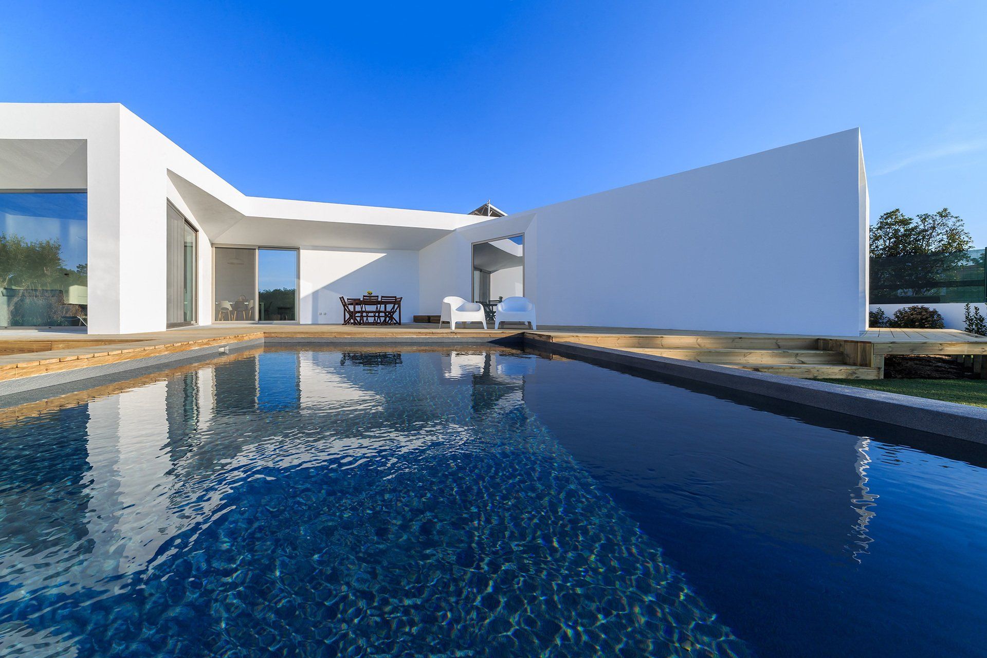Una moderna casa minimalista blanca con grandes ventanales se alza detrás de una piscina rectangular de color azul oscuro bajo un cielo despejado.