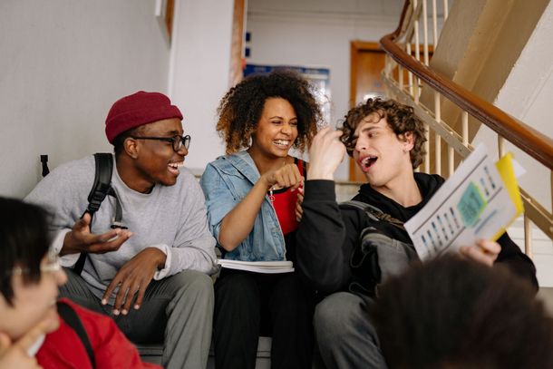 Students on stairs laughing, with papers. One wears a maroon beanie.