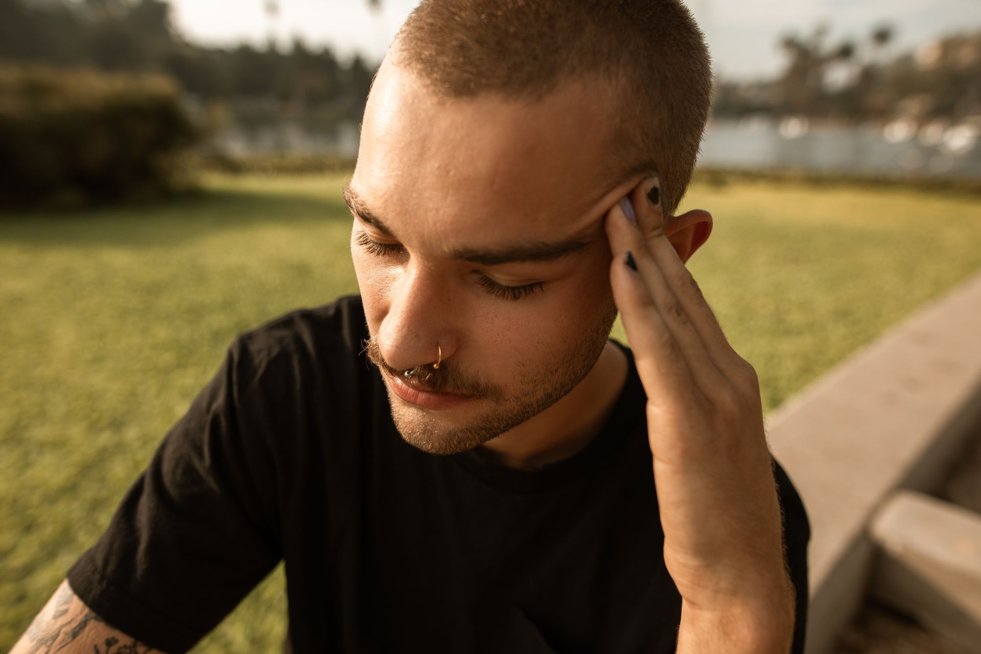 Man with short hair, nose ring, and painted nails, holding his head in a park.