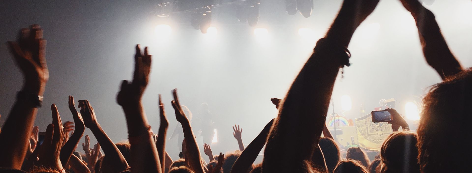 People raising their hands in the air at a concert. Stage lights shine through the smoke.