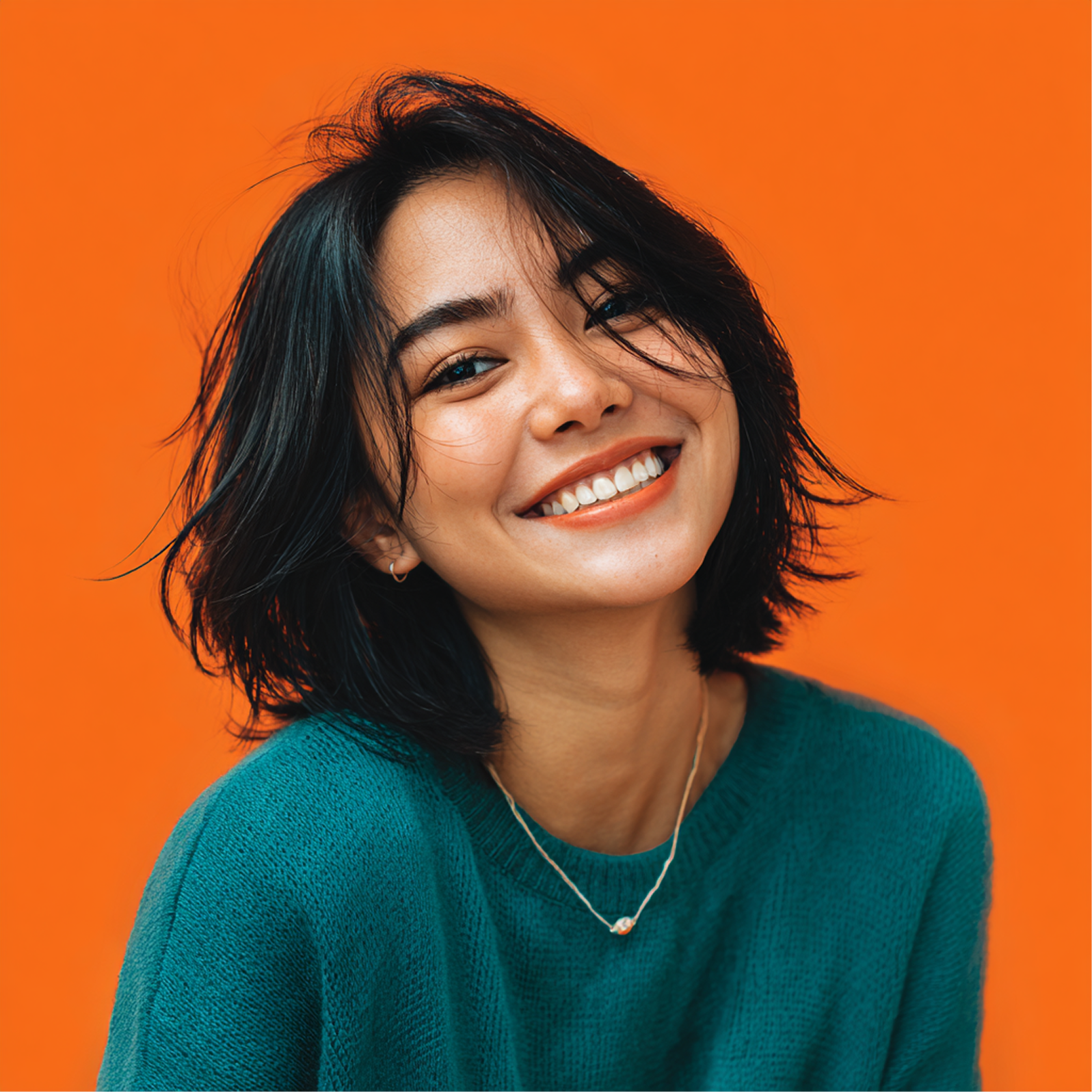 Woman with short, dark hair smiles broadly against an orange background. She wears a teal sweater.