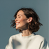 Smiling person with short dark hair in a white sweater, lit by sunlight against a blue-gray background