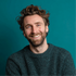 Smiling man with curly hair and beard in a dark sweater against a teal background