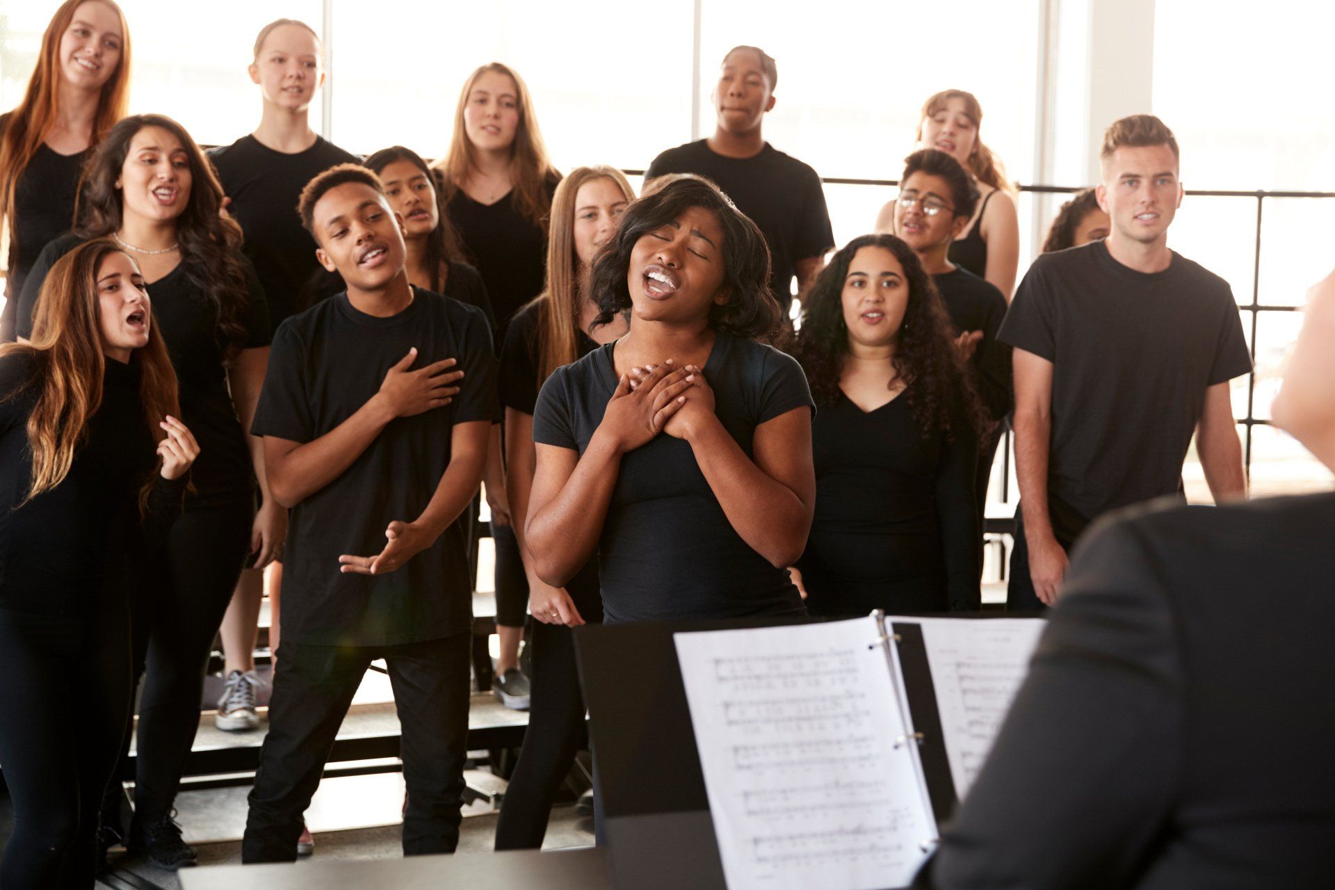 A choir of singers in black stands on risers, singing toward a conductor at a piano.