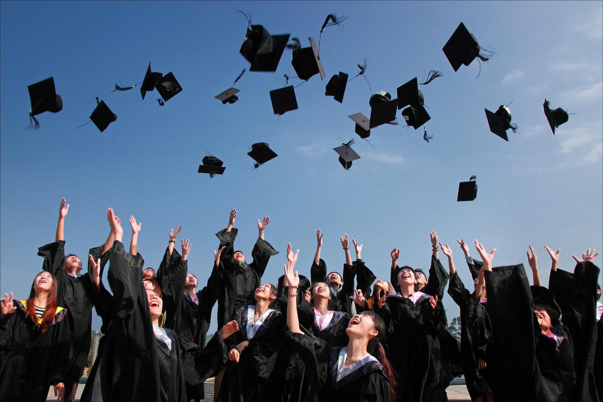 Graduates in black caps and gowns toss their mortarboards into a blue sky outdoors