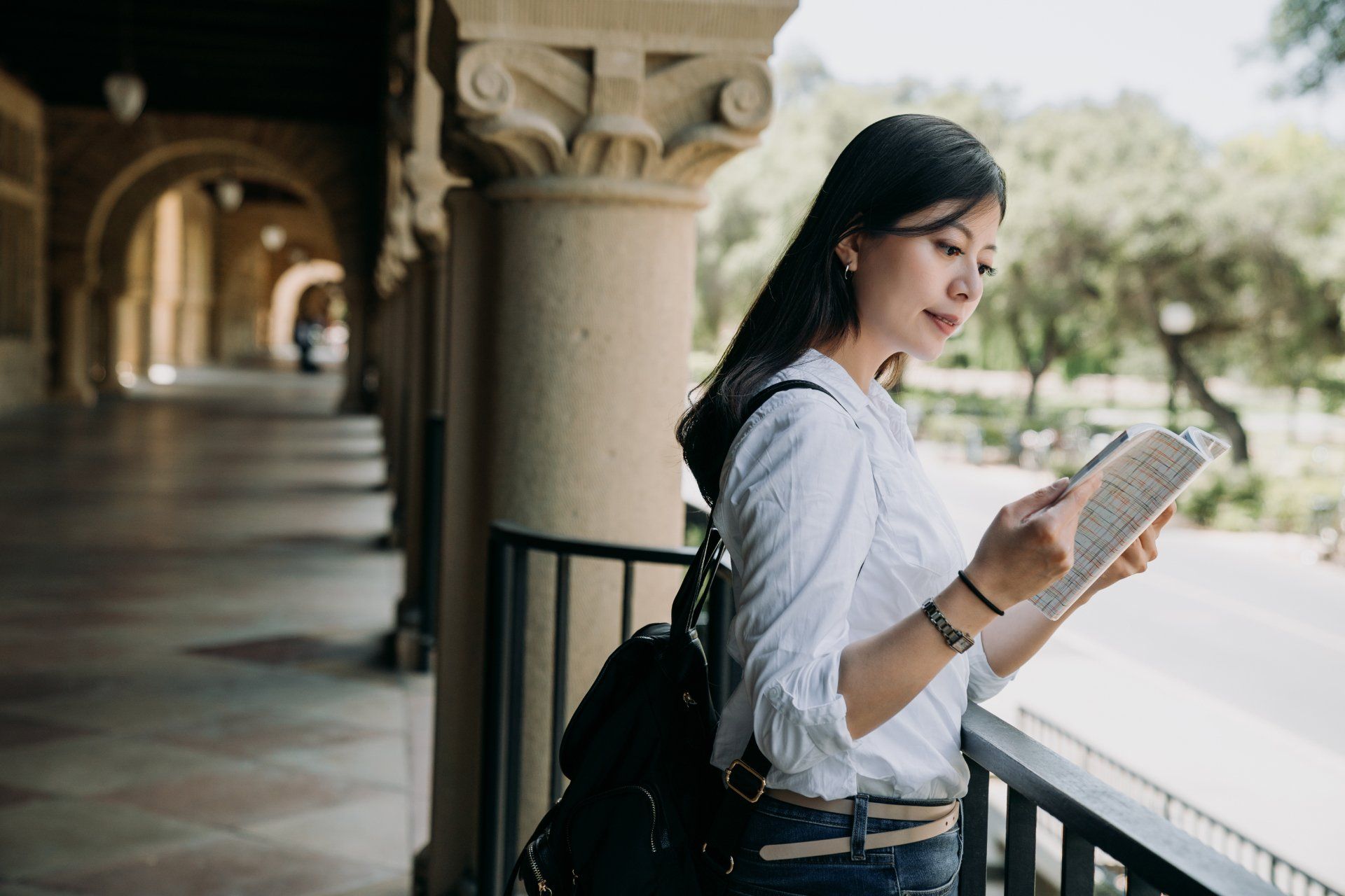 Woman reading book on a balcony with stone columns and a black backpack.
