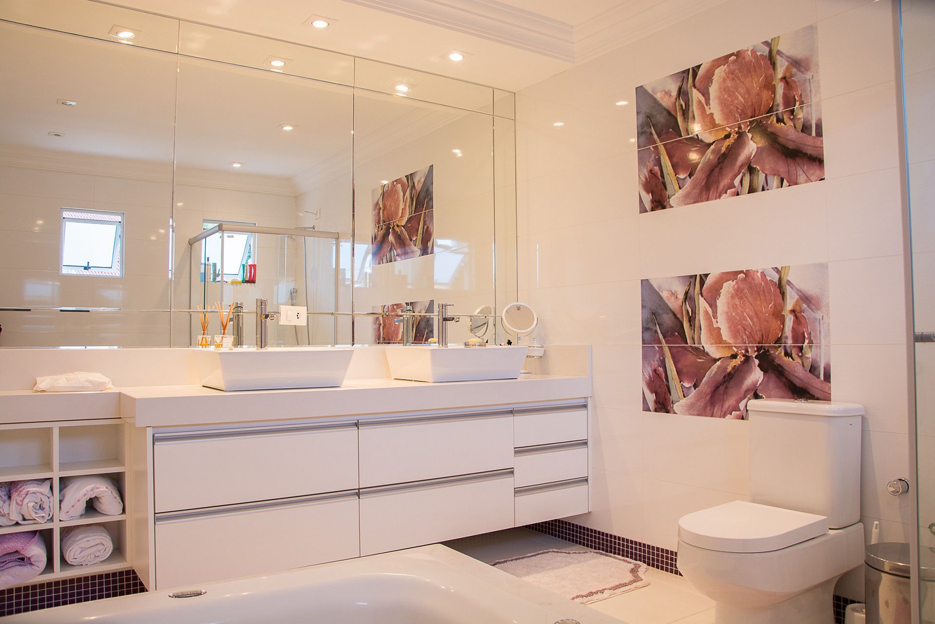Modern white bathroom featuring a dual-sink vanity, large wall mirror, toilet, and decorative floral wall art.