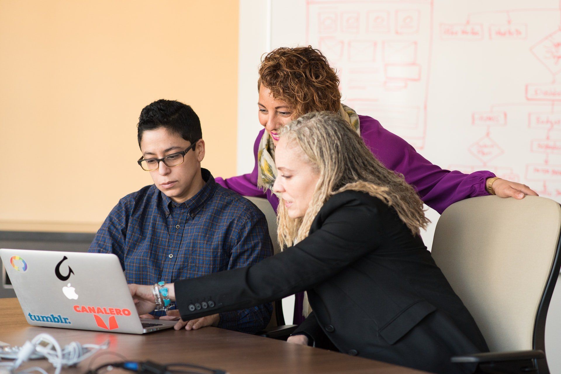 Three people collaborating at a table with a laptop; one points at the screen.