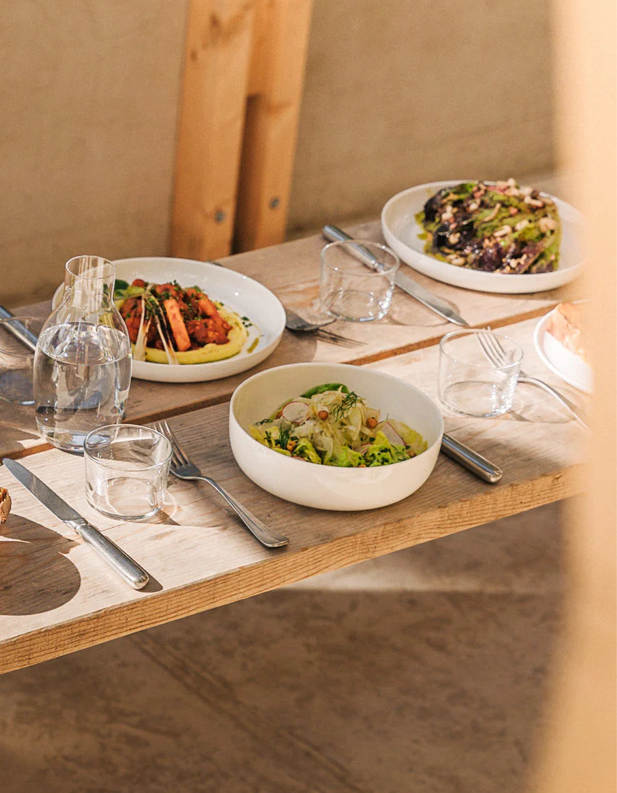 Wooden table set with dishes of food, glasses, and silverware. Sunlight streams in.