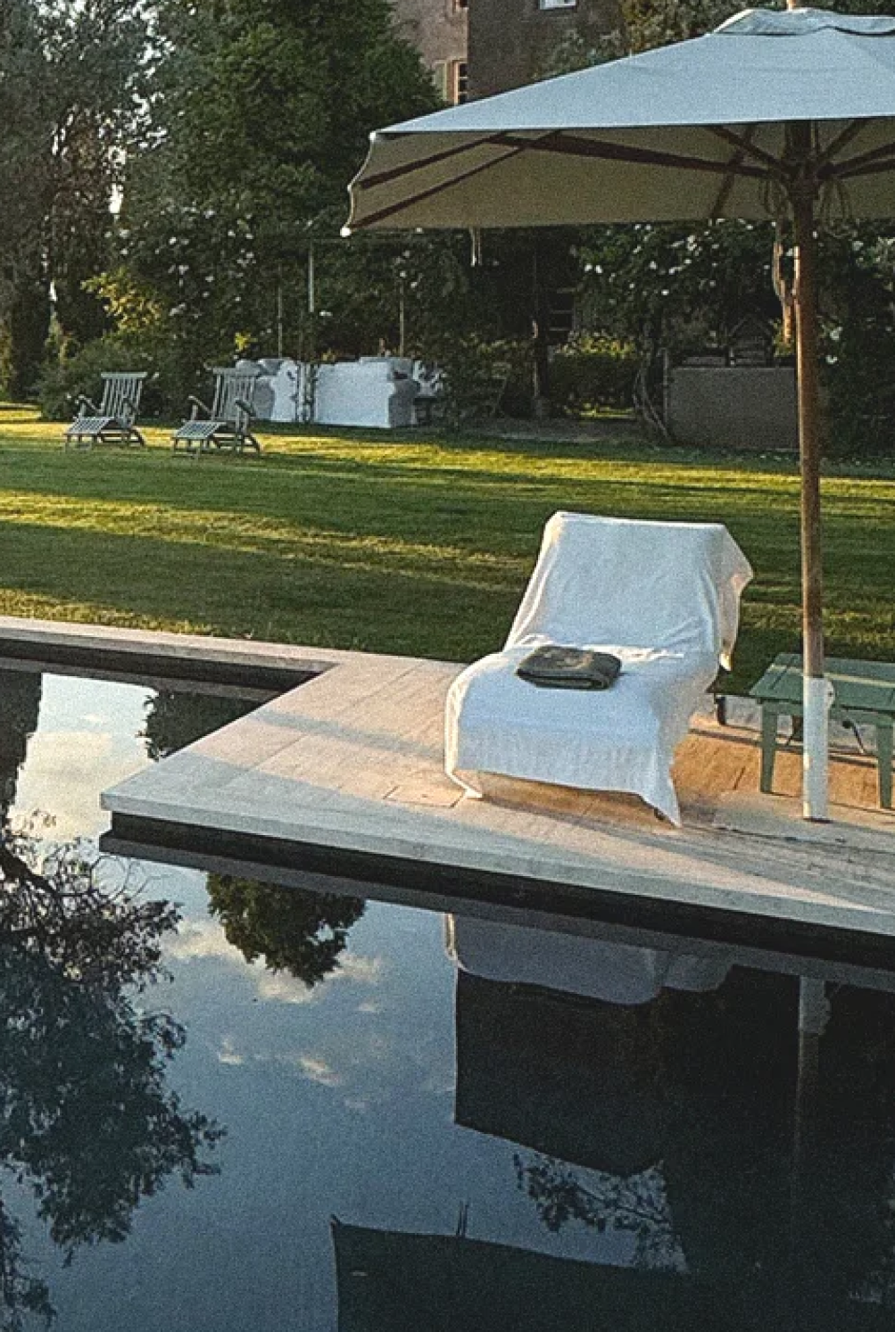 A pool with a deck and a chaise lounge covered in white, an umbrella, and a green lawn.
