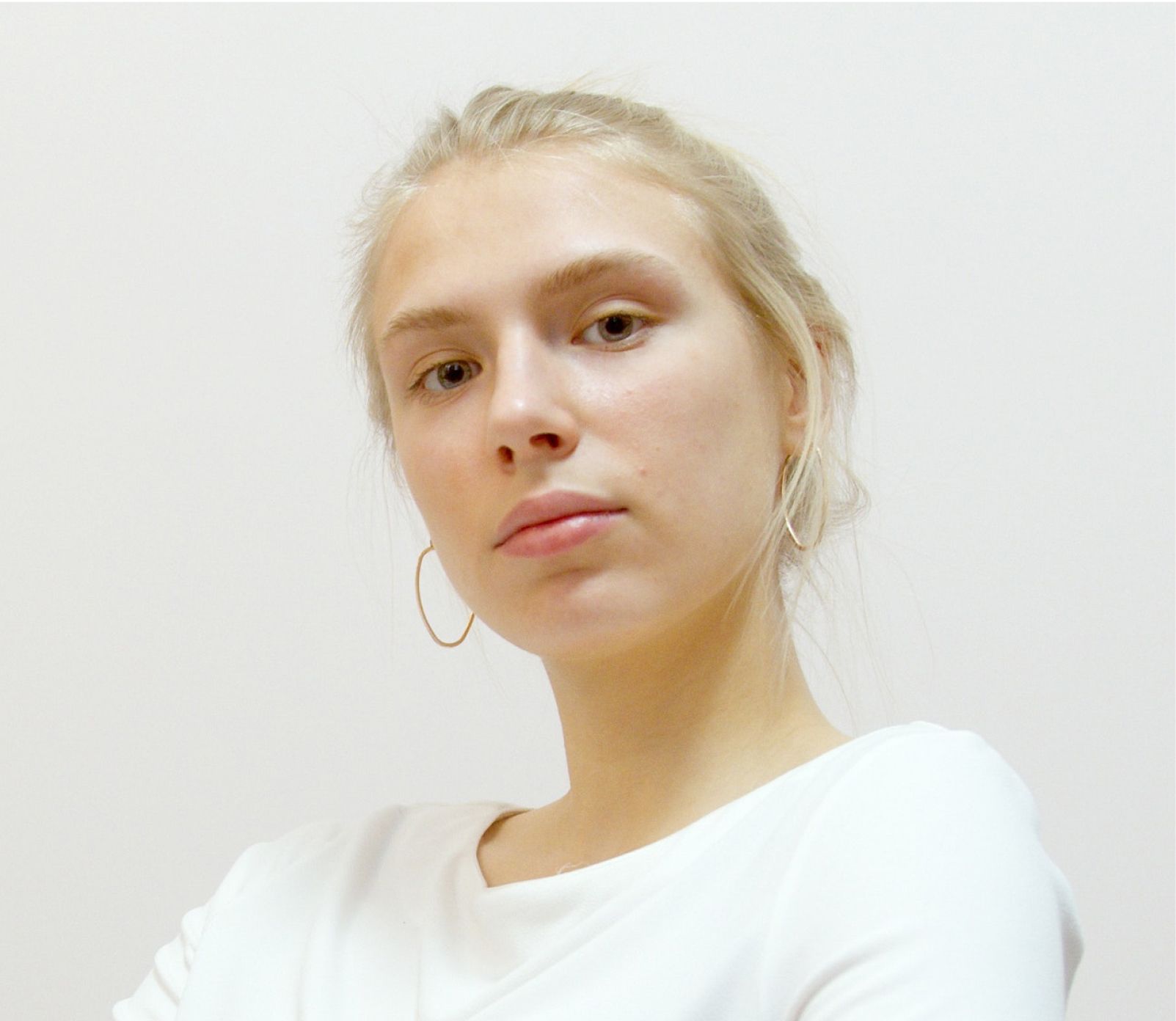 Blonde woman in a white shirt looks directly at the camera. She has simple hoop earrings and a neutral expression.