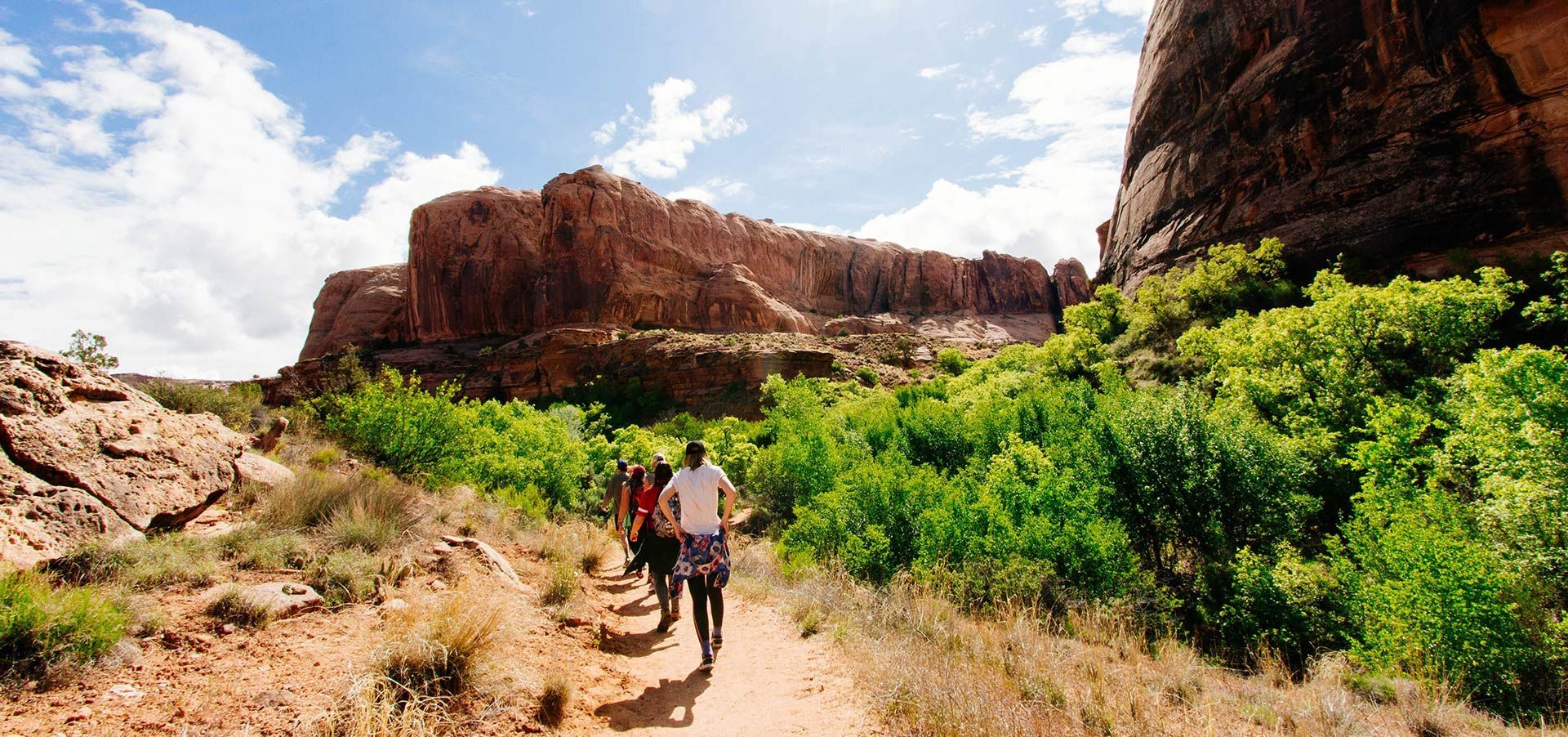 People walking on a dirt path with rock formations and green vegetation on either side. Blue sky with clouds.