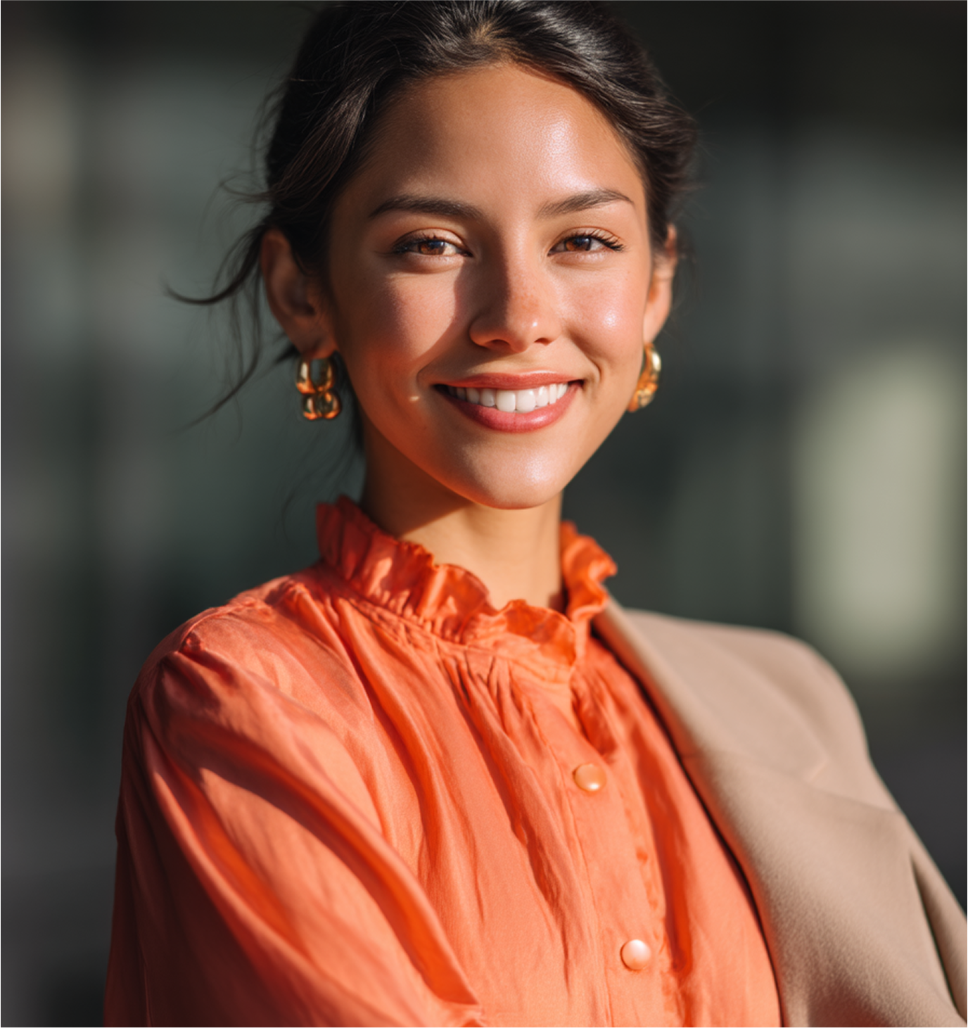 Smiling person in an orange blouse and tan blazer against a softly blurred background