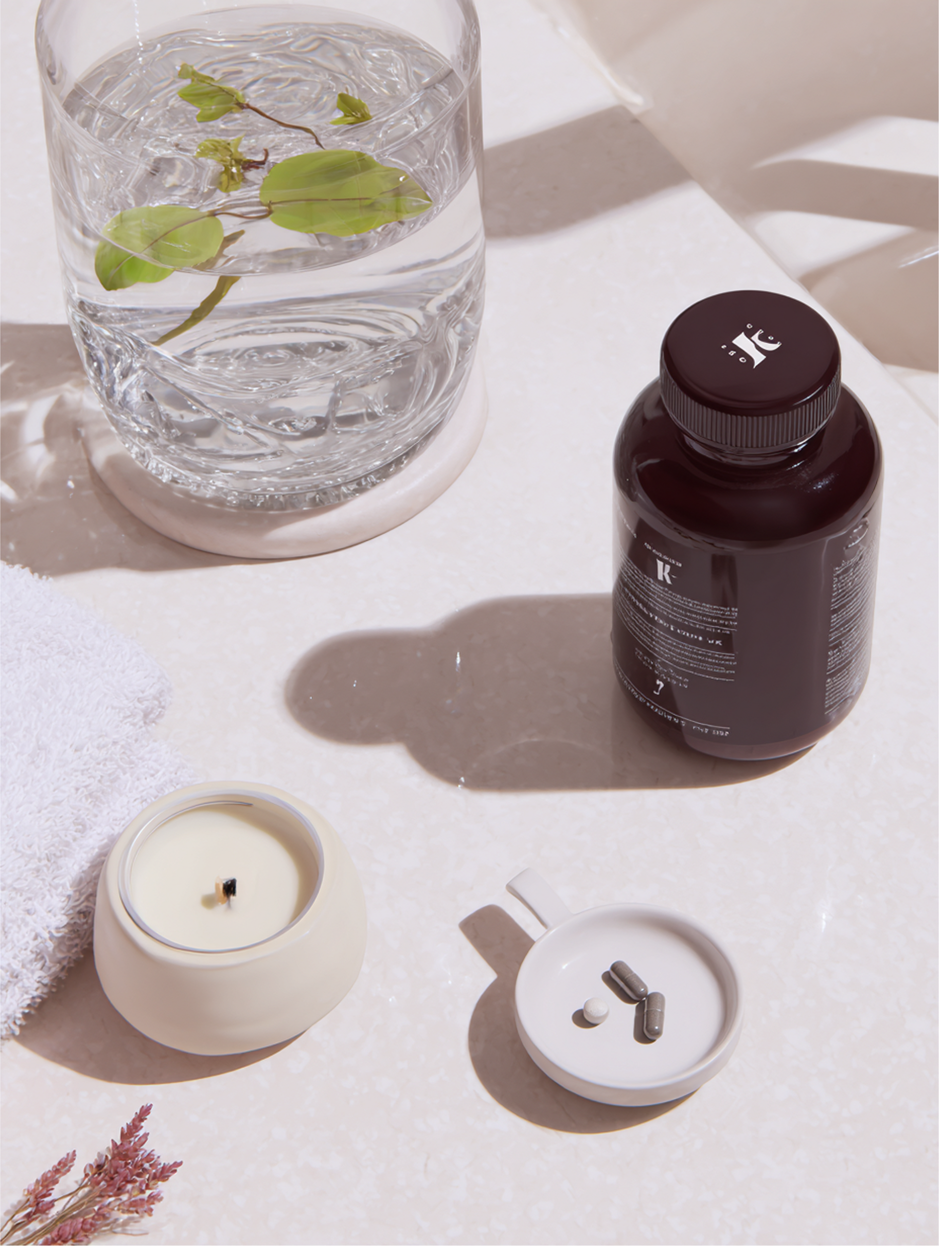 Glass of water with leaves, supplement bottle, candle, and towel on a white surface.