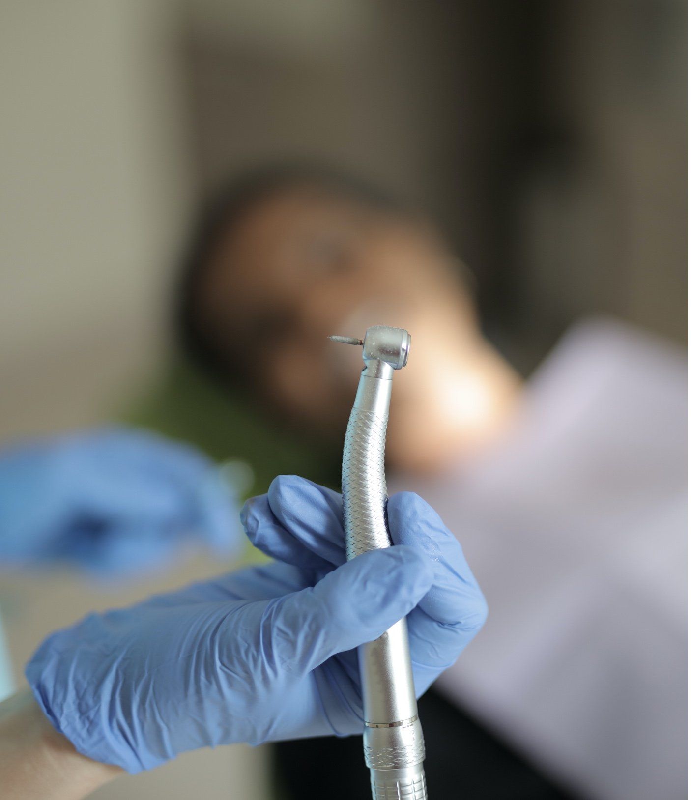 Hand in blue glove holding dental drill, patient blurred in background.