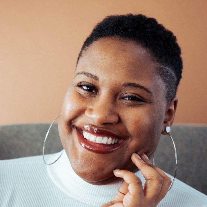 Woman with short, dark hair, wearing large hoop earrings and a white turtleneck smiles, resting a finger on her cheek.