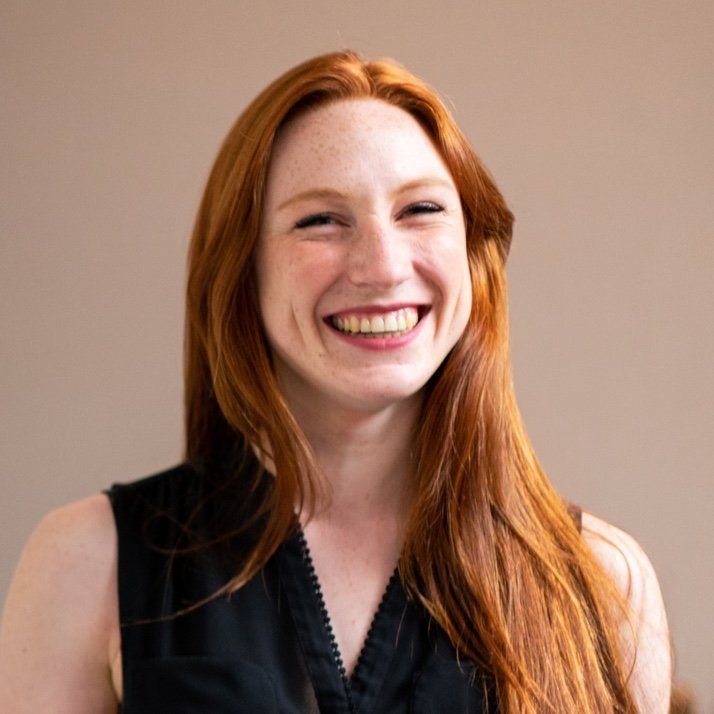 Woman with long red hair, smiling broadly in a dark top against a neutral background.