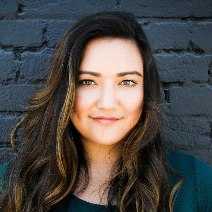 Woman with long, wavy brown hair, smiling, in front of a dark brick wall.