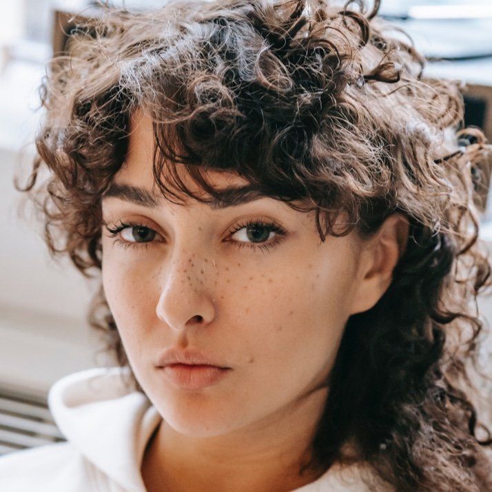 Woman with curly hair and freckles, looking directly at the camera with a neutral expression.