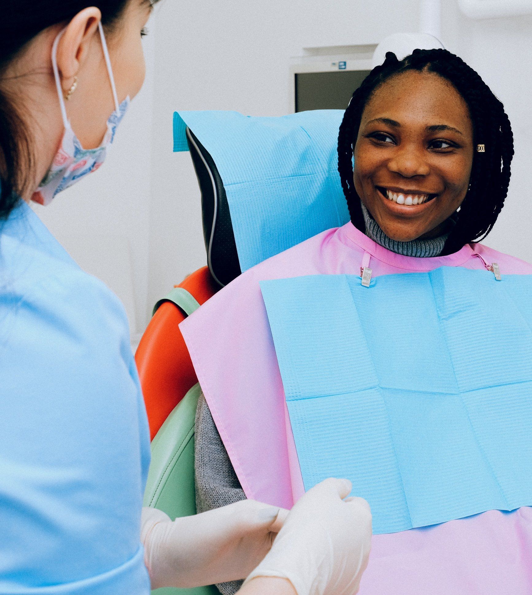 Dentist with mask adjusts patient's bib. Patient smiles in dental chair.