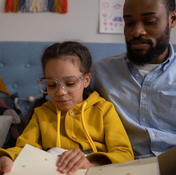 Adult reading with child in yellow hoodie and glasses on a couch.