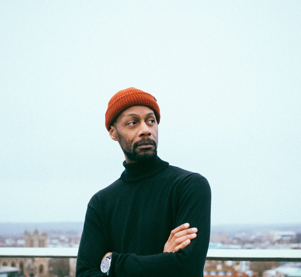 A man wearing a beanie and a black sweater is standing with his arms crossed.