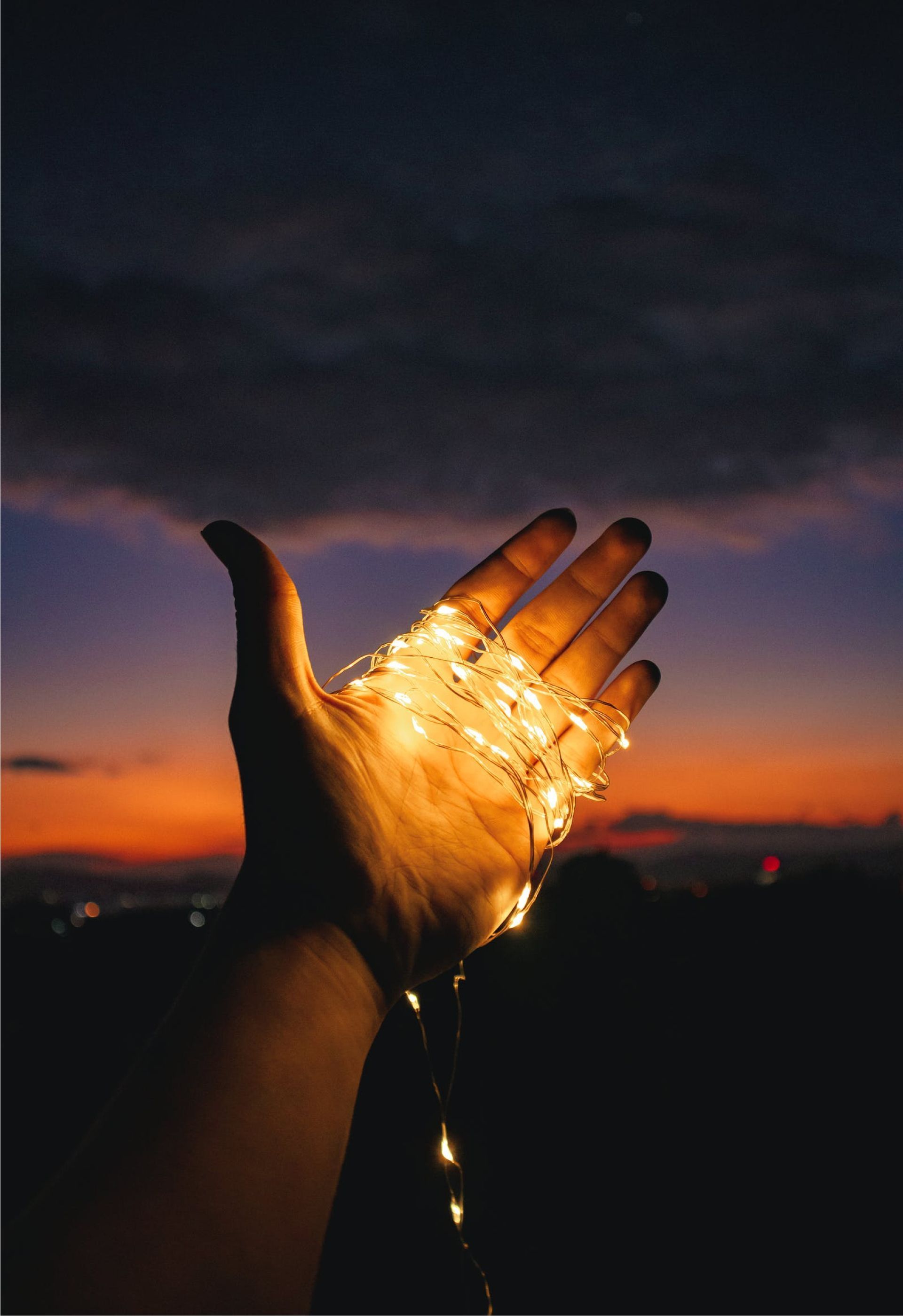 Hand holding string lights, set against a dark, colorful sunset sky.
