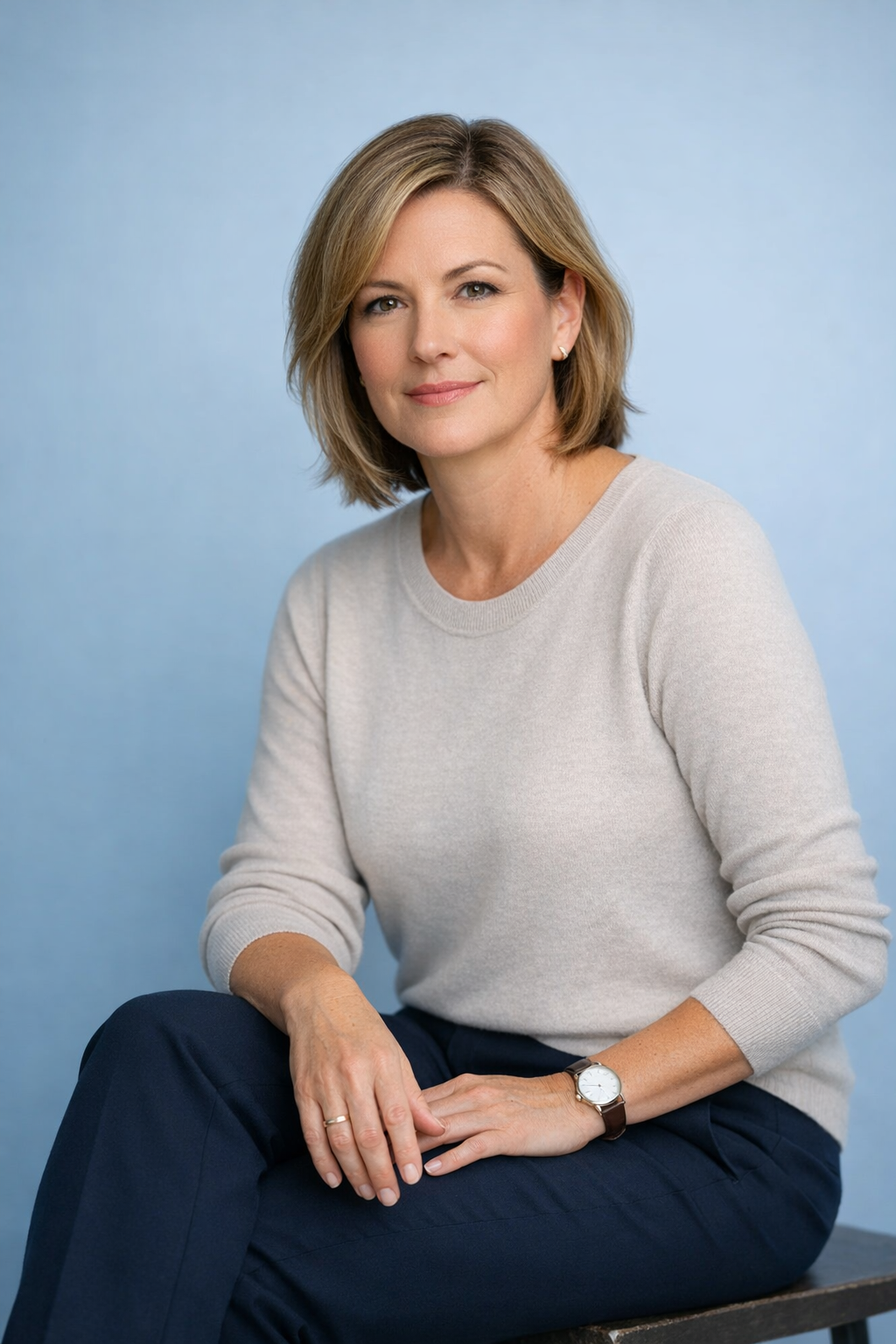 Seated woman in a light sweater and dark pants, posing against a blue studio backdrop.