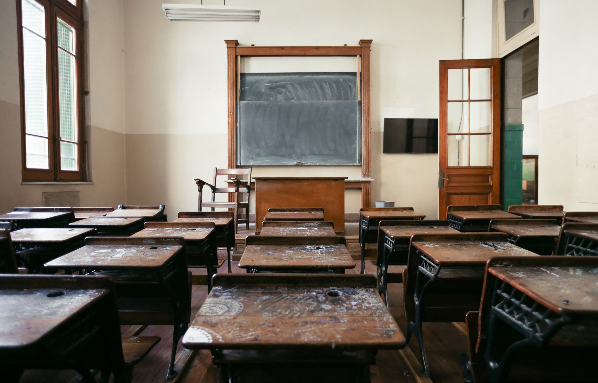 Empty classroom with old wooden desks facing a chalkboard. Natural light streams in.