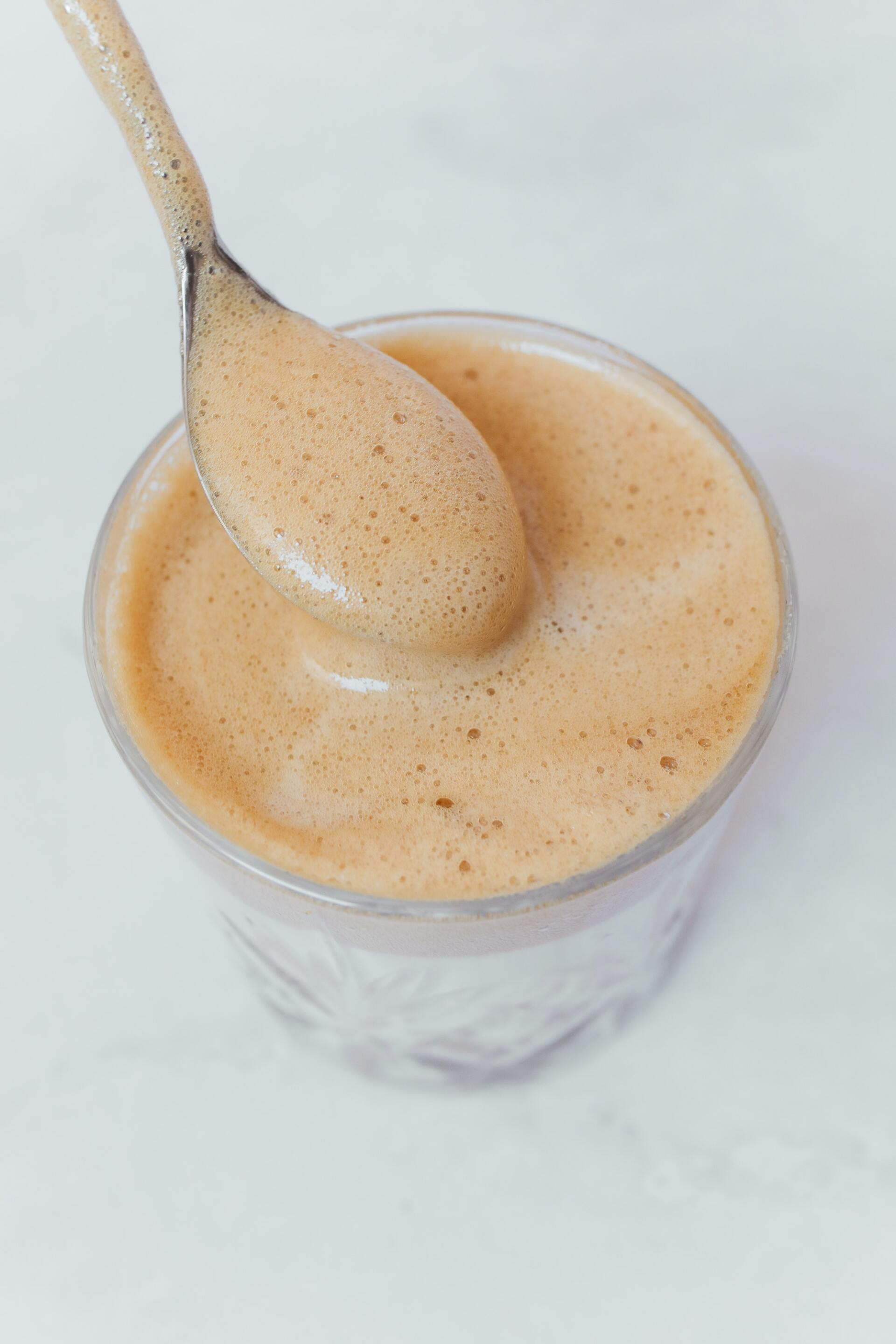 A spoon scoops up coffee foam from a glass; creamy brown foam contrasts with clear liquid and white marble.