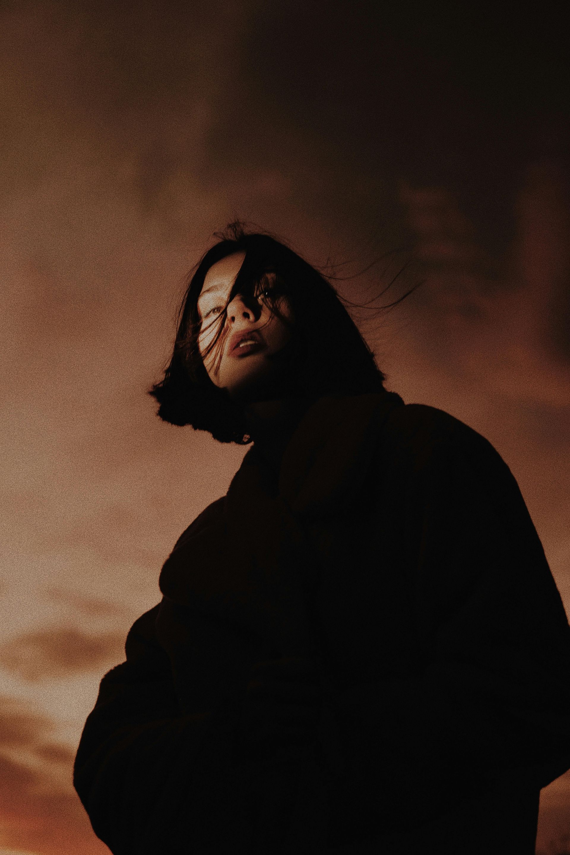 A woman wearing glasses and a black jacket is standing in front of a cloudy sky.