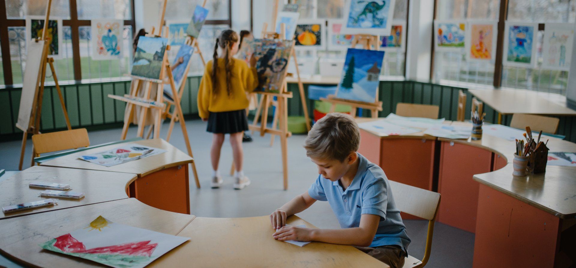 Children in an art class: One boy draws at a table while another girl paints on an easel.