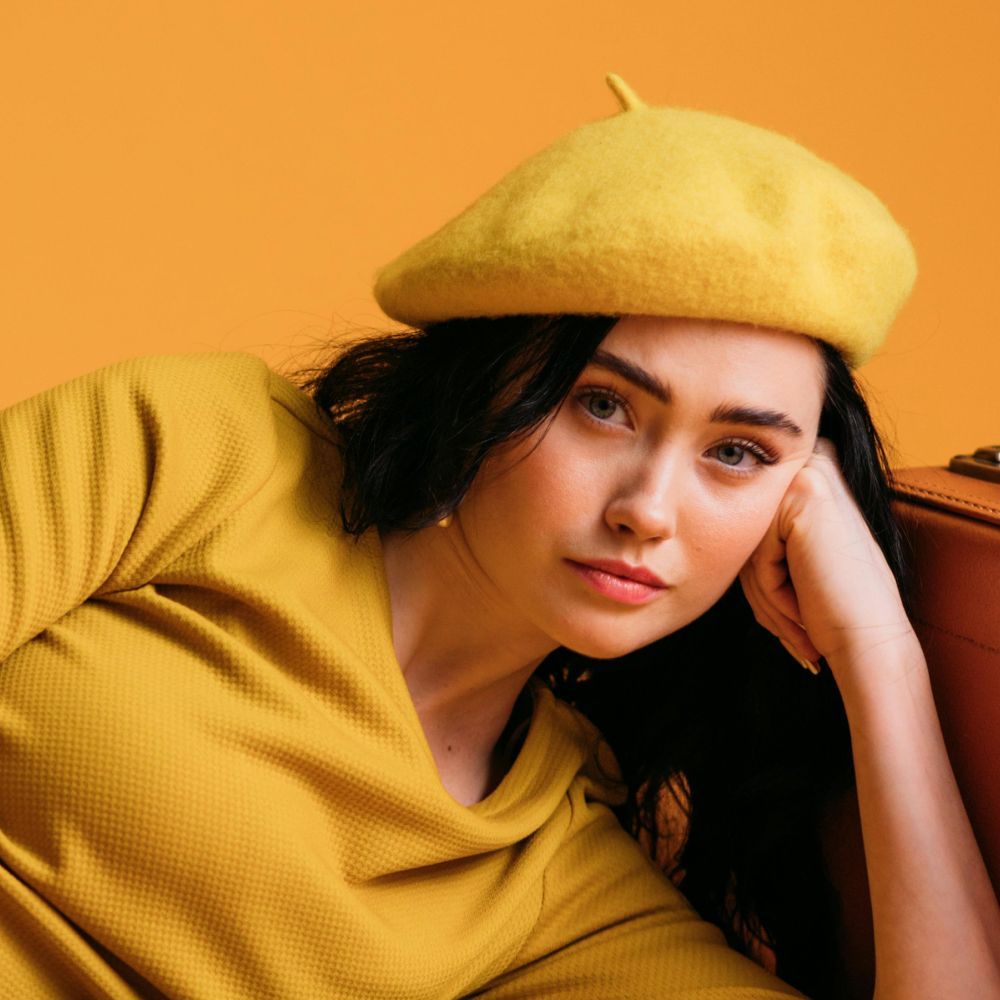 Woman wearing yellow beret and shirt, leaning against a suitcase, with an orange backdrop.