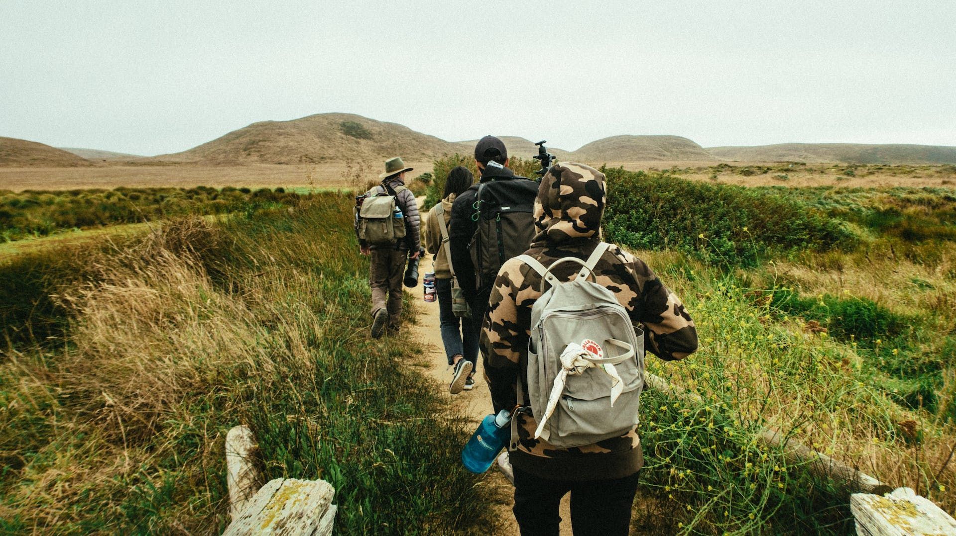 A group of people wearing backpacks and hiking clothes walks along a dirt path through a grassy, arid landscape.