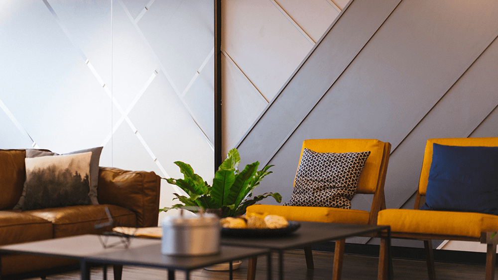 A modern living room with brown couch, yellow chairs, black table, and a potted plant in front of a textured gray wall.