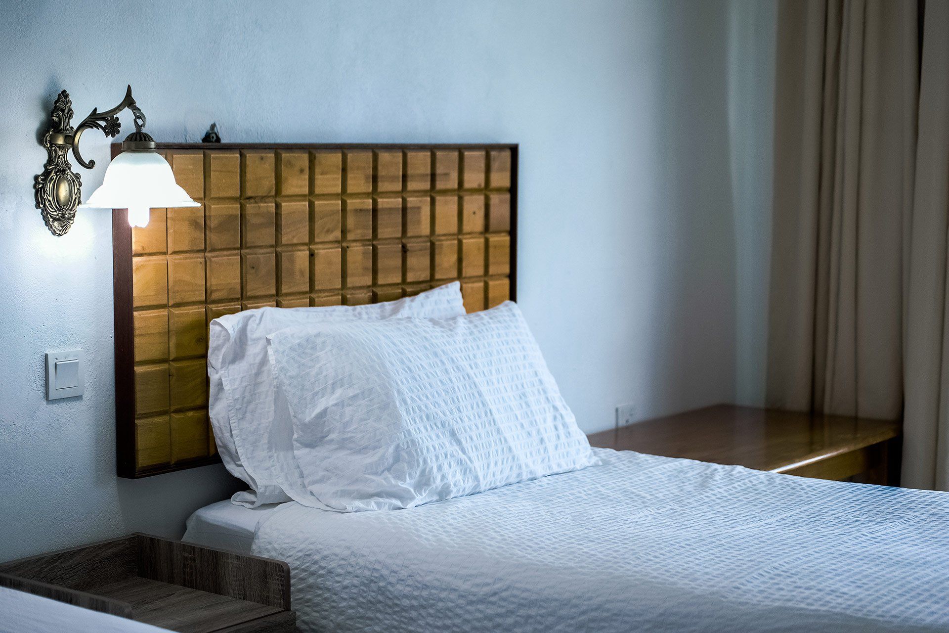 Bed with a white duvet and pillow, wooden headboard, and a sconce providing light.