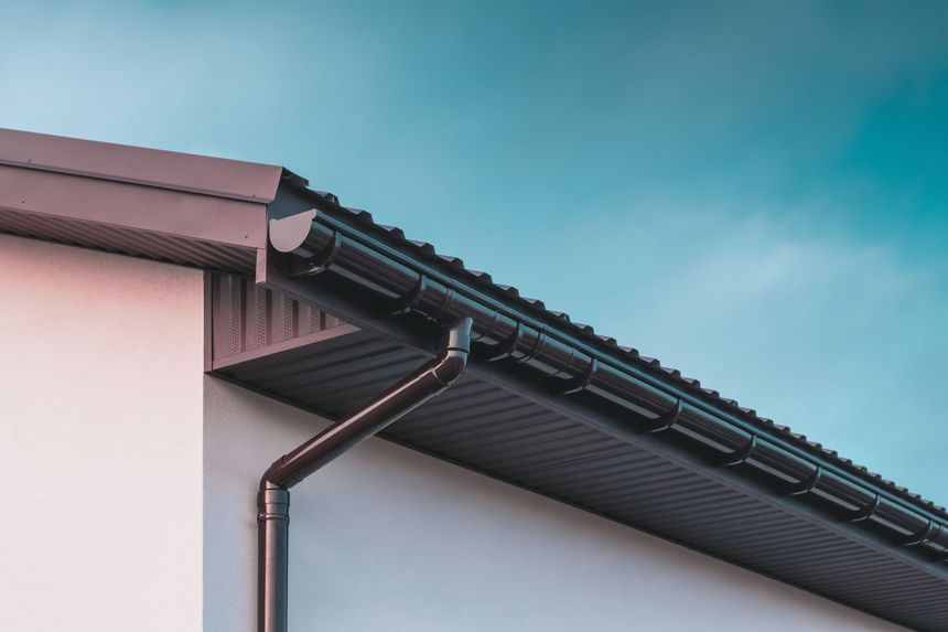 Close-up of a house roof with dark gutters and downspout against a blue sky