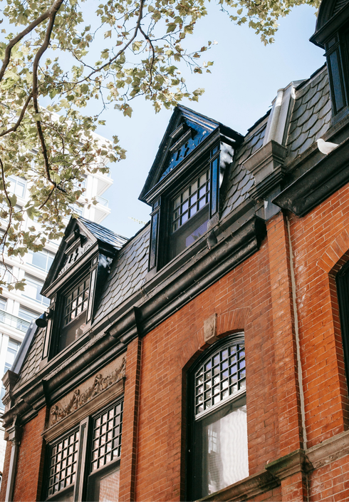 Red brick building with dark roof dormers and arched windows beneath tree branches